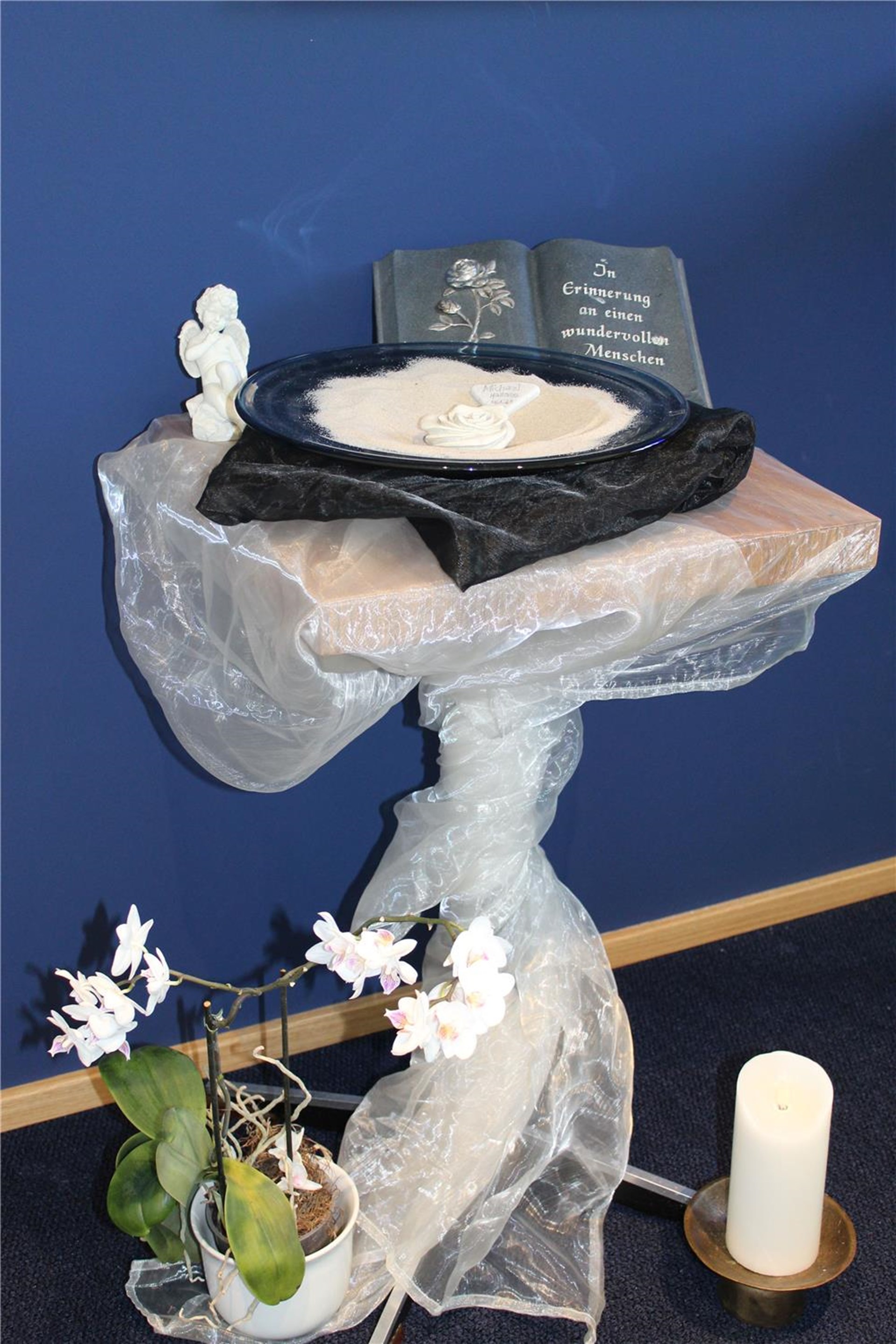 Ein Tisch ist in eine Gardiene eingehüllt. Auf ihm stehen eine Schale, eine Erinnerungsanzeige und Kerzen.